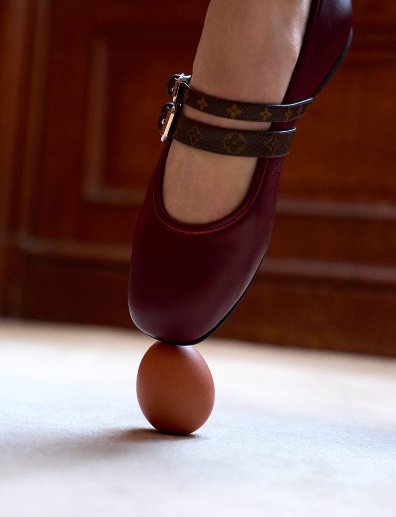 Close-up of a woman's burgundy shoe with patterned straps crushing a brown egg on a beige surface against a wood background.