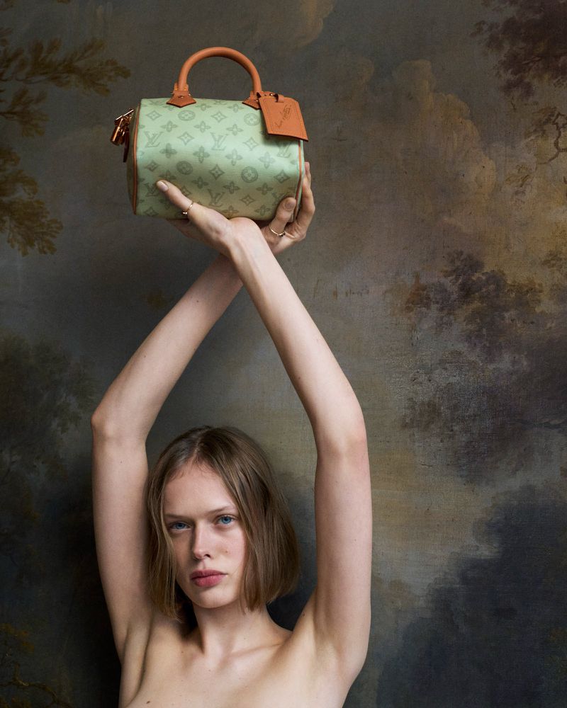A fair-skinned young adult with short blonde hair poses in front of an artistic background holding a light green monogrammed handbag.
