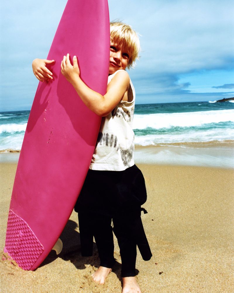 Young boy hugs a pink surfboard on a sunny beach with ocean waves and blue sky in the background, wearing a patterned shirt and wetsuit pants.