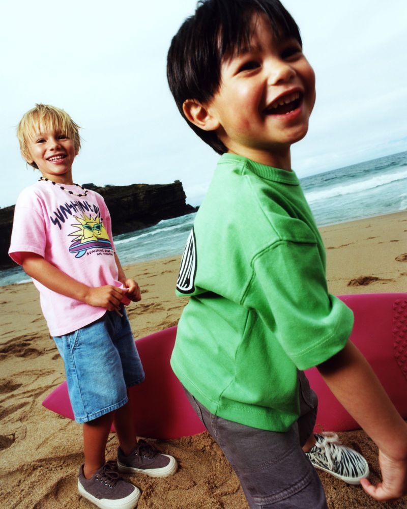 Two young boys smile brightly on a sunny beach with a surfboard. One wears a green shirt, the other a pink shirt with blue shorts.