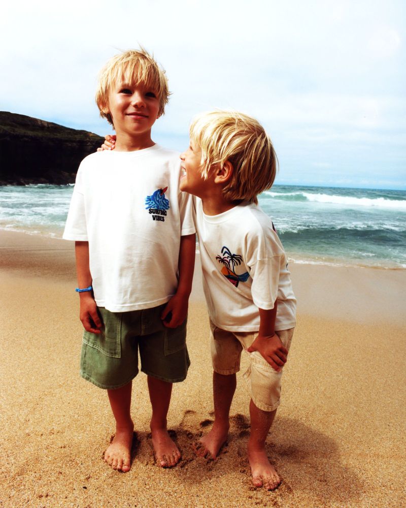 Two young boys pose on a sandy beach with ocean waves and a distant green hillside in the background, both wearing light-colored shirts and shorts.
