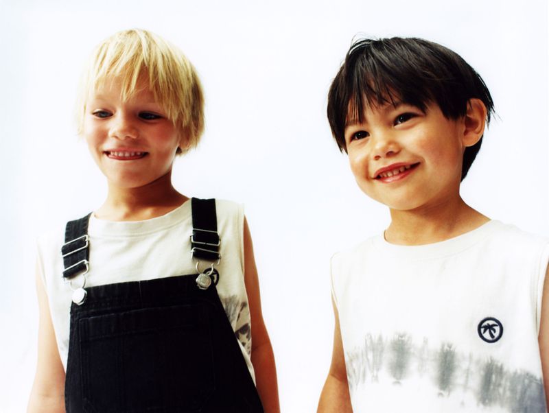 Two young children, one with blonde hair in overalls and another with dark hair in a graphic tee, smile against a white backdrop.
