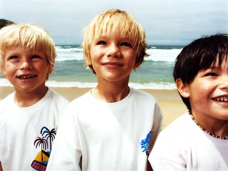 Three young boys stand on a beach with blond and dark hair, wearing matching white t-shirts. Ocean waves and a sandy beach are visible.