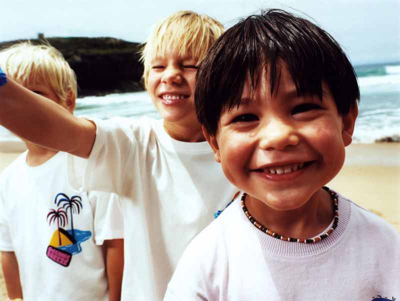 Three smiling children on a beach wearing white t-shirts. The foreground child wears a beaded necklace. Ocean and green hillside in background.
