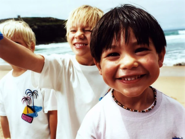 Three smiling children on a beach wearing white t-shirts. The foreground child wears a beaded necklace. Ocean and green hillside in background.