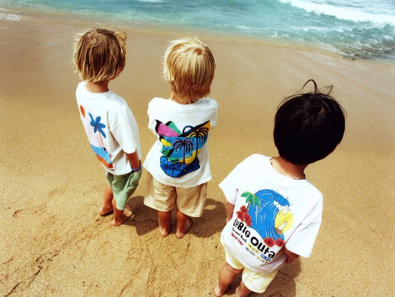 Three children stand on a sandy beach facing the ocean waves. They wear matching t-shirts with colorful tropical designs on their backs.