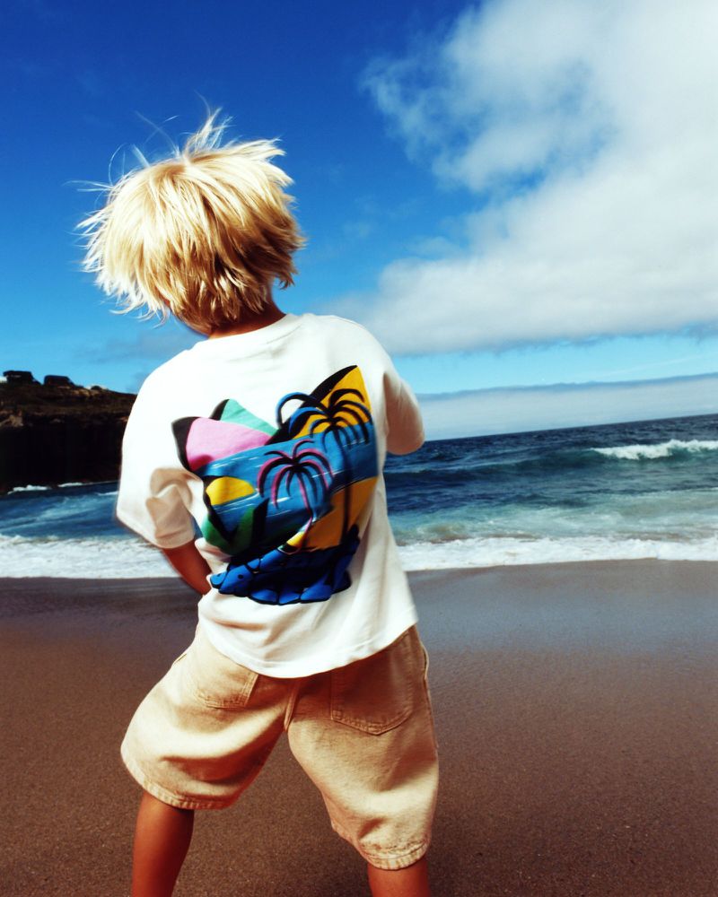 Young child stands on a beach with the ocean and sky in the background, wearing a white t-shirt and khaki shorts on a sunny day.