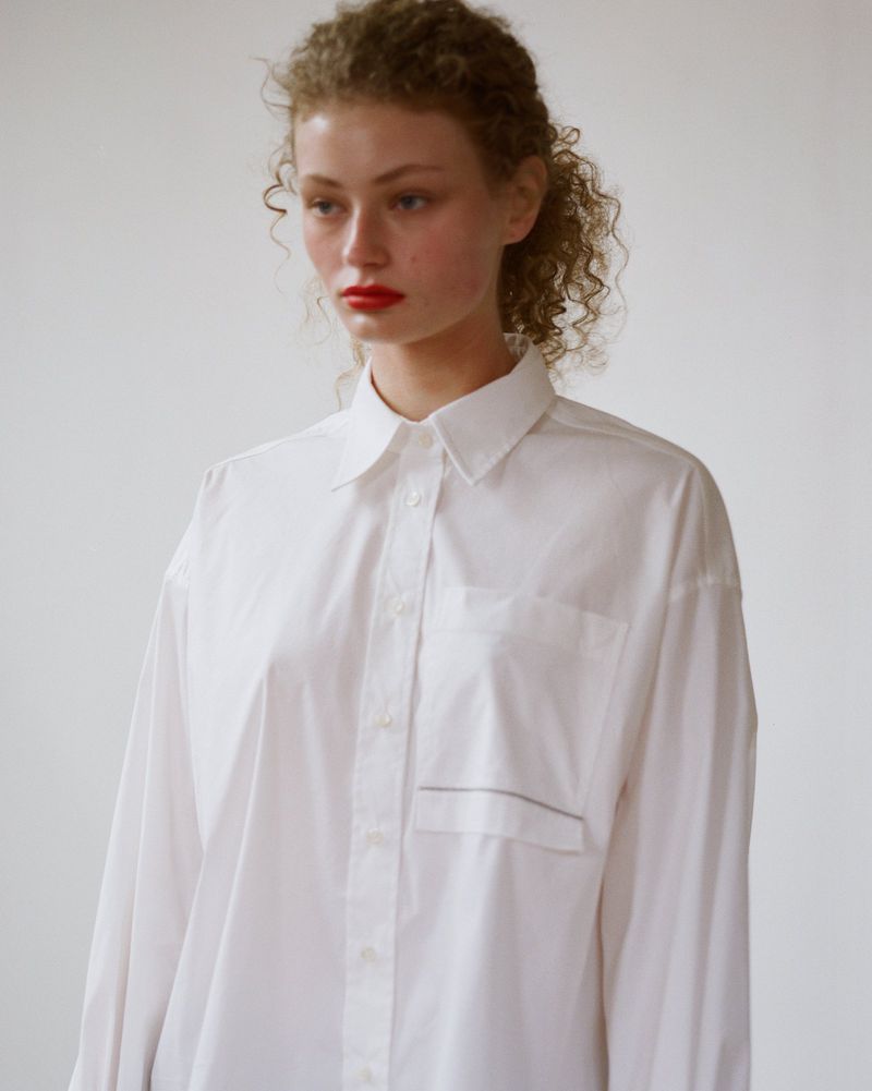 Portrait of a woman with curly hair wearing an oversized white button-down shirt, red lipstick, and a neutral background.
