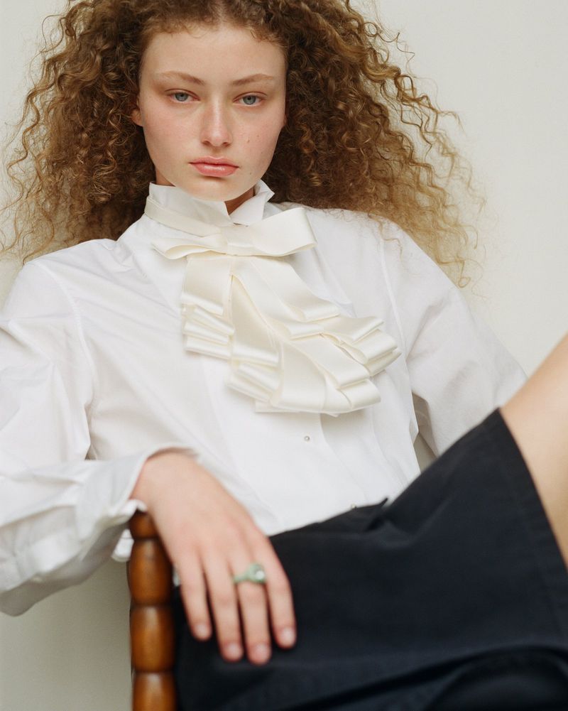 A young woman with curly hair poses indoors wearing a white collared shirt with a ruffled bow, captured in a medium shot.
