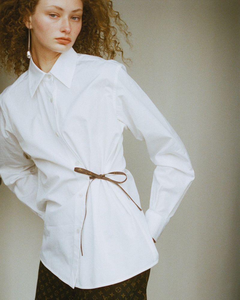 Female model in a white button-down shirt with a brown ribbon belt, posing in front of a plain background with neutral colours.
