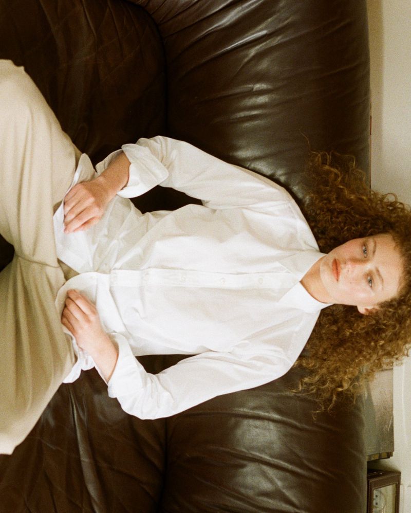 Woman with curly hair lying on a dark leather couch, wearing a white shirt and light-colored pants in a relaxed pose.