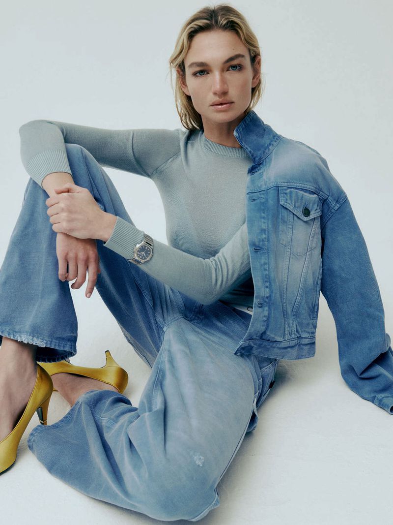 A blonde woman in a light blue sweater and jeans sits on a white background with a denim jacket and yellow heels in a fashion shot.