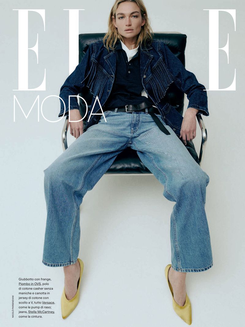 Fashion editorial shot of a model in denim jacket and wide leg jeans, seated on a chair in a bright studio setting, magazine cover.