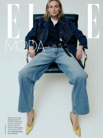 Fashion editorial shot of a model in denim jacket and wide leg jeans, seated on a chair in a bright studio setting, magazine cover.