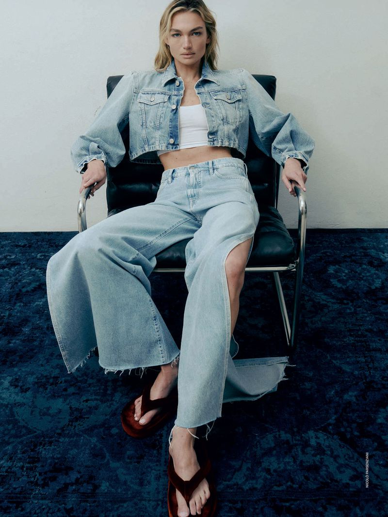 A woman in a light blue denim jacket, wide-leg jeans with a slit, and brown sandals poses in a black chair on a blue carpet.