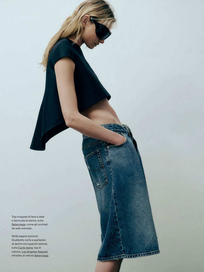 Fashion editorial shot: blonde woman in a cropped top and denim culottes poses against a plain wall, showcasing minimalist style.
