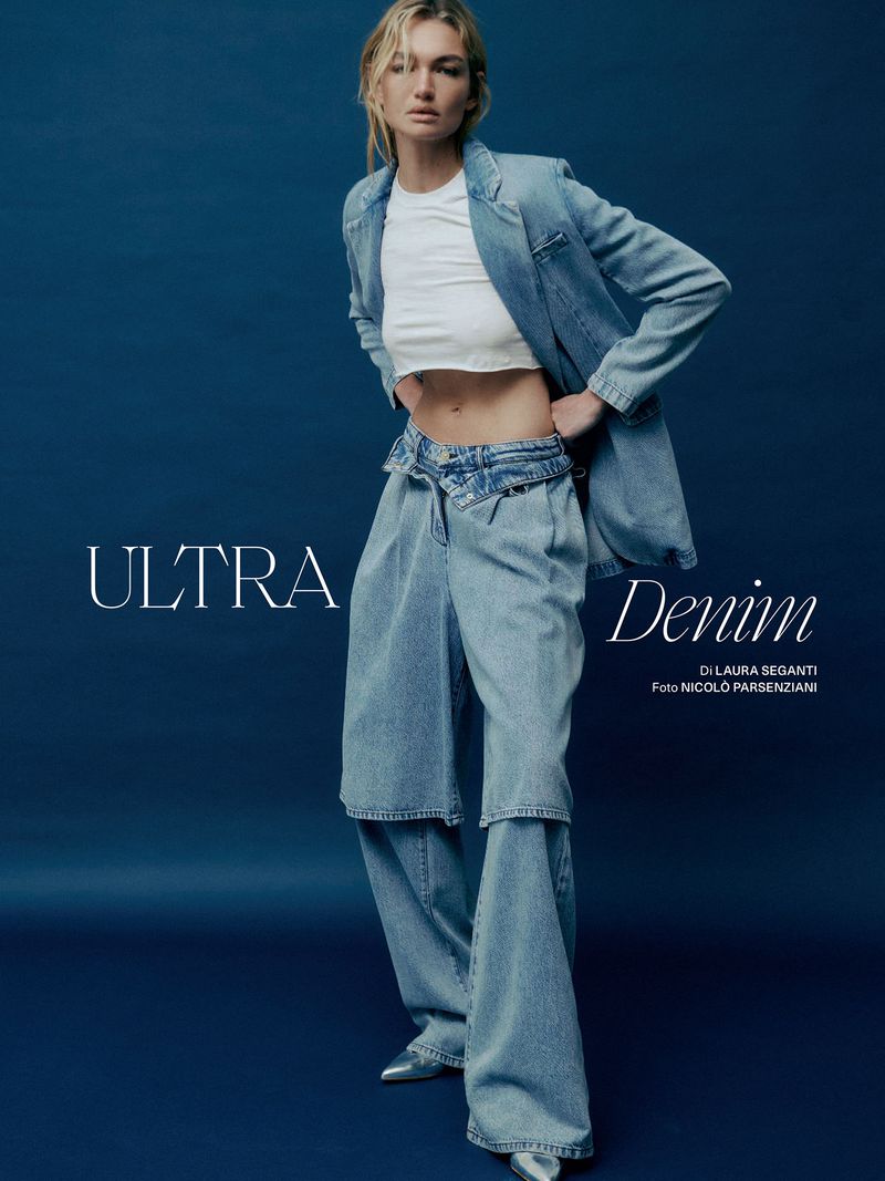 Fashion shot of a woman in a light-blue denim jacket and jeans, standing in front of a dark blue background in a studio environment.