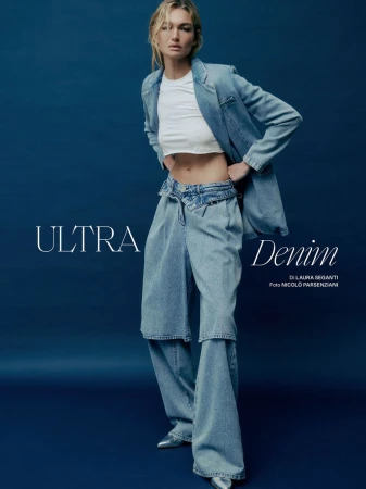 Fashion shot of a woman in a light-blue denim jacket and jeans, standing in front of a dark blue background in a studio environment.