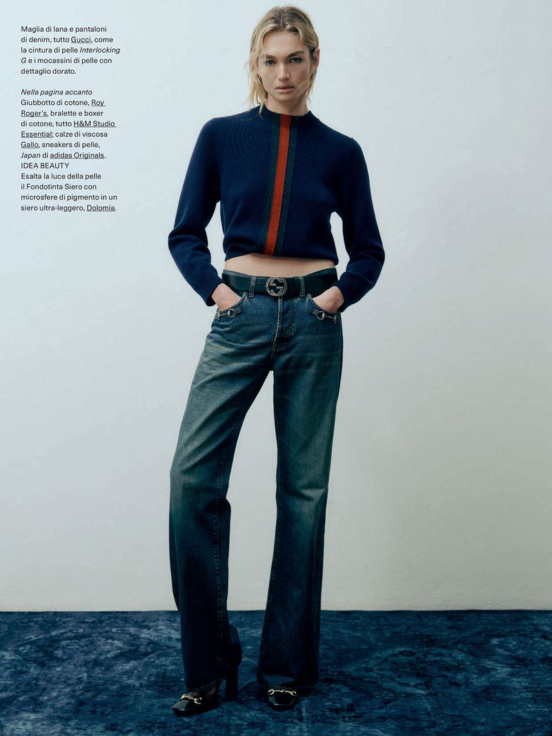 Fashion editorial shot of a fair-skinned female model with blond hair wearing a cropped blue sweater and jeans in a studio.