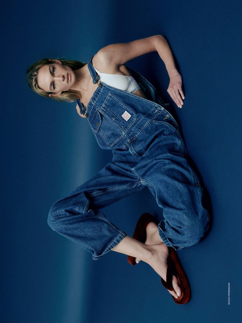 Female fashion model in denim overalls and a white top strikes a pose against a solid dark blue backdrop in a studio setting.