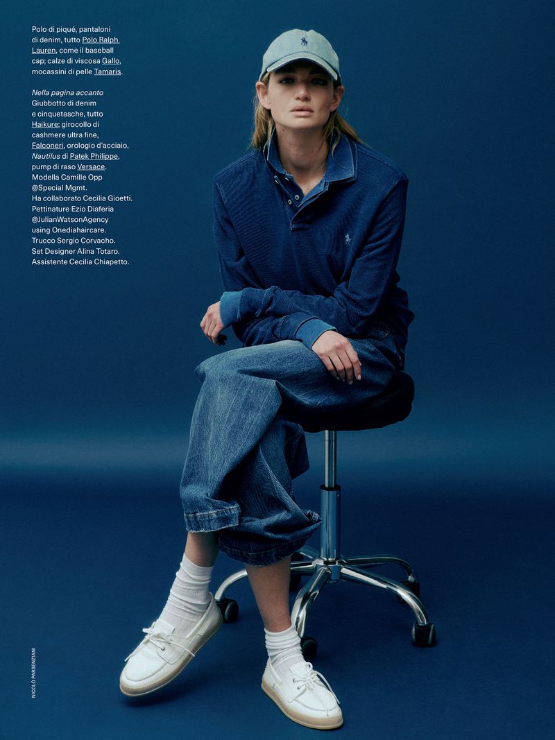 Adult female fashion model posing seated on a chrome chair in blue denim polo shirt and jeans with white shoes against blue backdrop.