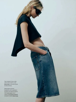 Model wearing dark sunglasses, dark cropped top, and blue denim shorts standing against a grey background for Elle Italia