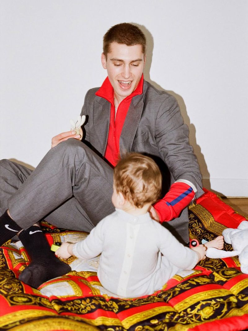 Young adult male in grey suit and red shirt sits with baby on patterned blanket, interacting with toys in bright indoor setting.