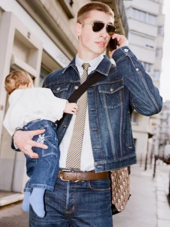 Busy parent walks down city street with baby, talking on phone, wearing denim jacket and sunglasses, with European architecture.