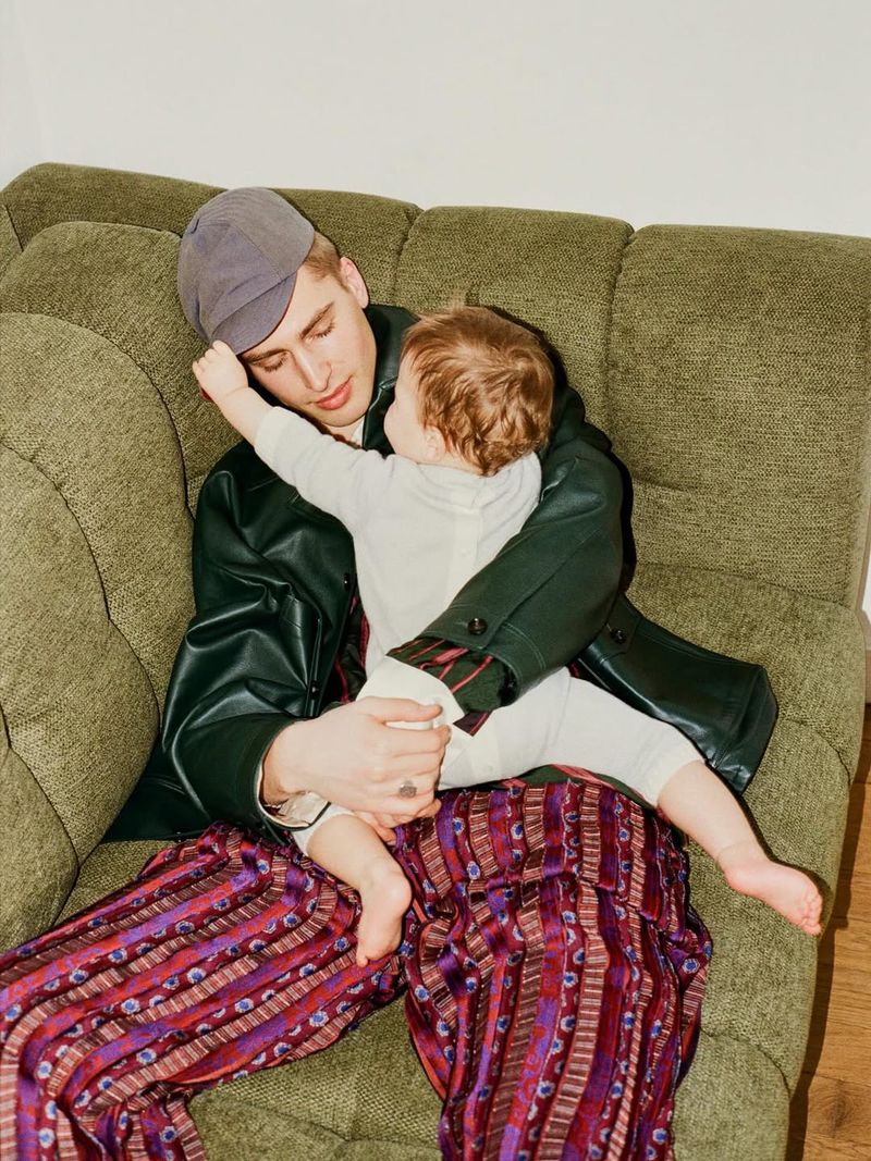 Man and toddler pose on green textured sofa. Young father embraces child in casual clothing. Indoor portrait with simple backdrop.