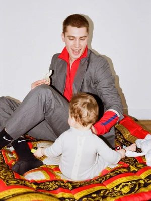 Man in a grey suit and red shirt seated with a child on a patterned mat against a white wall