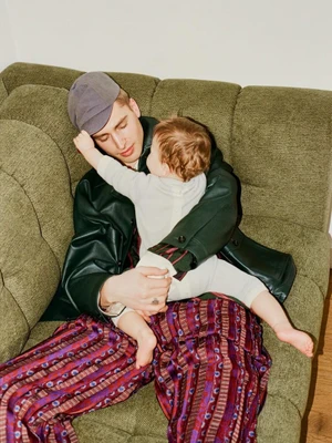 Man wearing a cap and dark jacket holding a baby on an olive green sofa for FAZ Magazine editorial