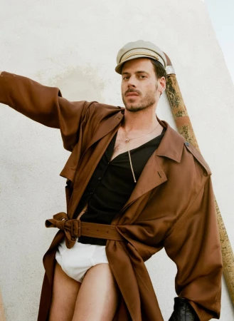 Stylish man in an open brown trench coat, black top, white underwear, and sailor cap poses against a textured wall, exuding confidence.