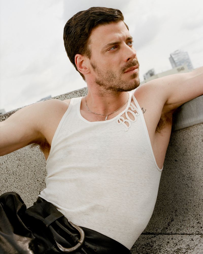 Male model poses outdoors in a white tank top and black leather pants, sitting on a concrete ledge against a blurred cityscape backdrop.