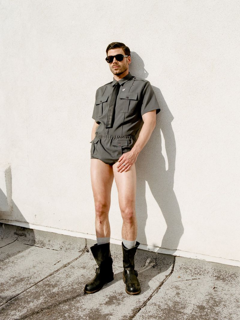 Full-length shot of a man posing in a short jumpsuit, boots, and sunglasses against a white wall outdoors with soft, diffused light.