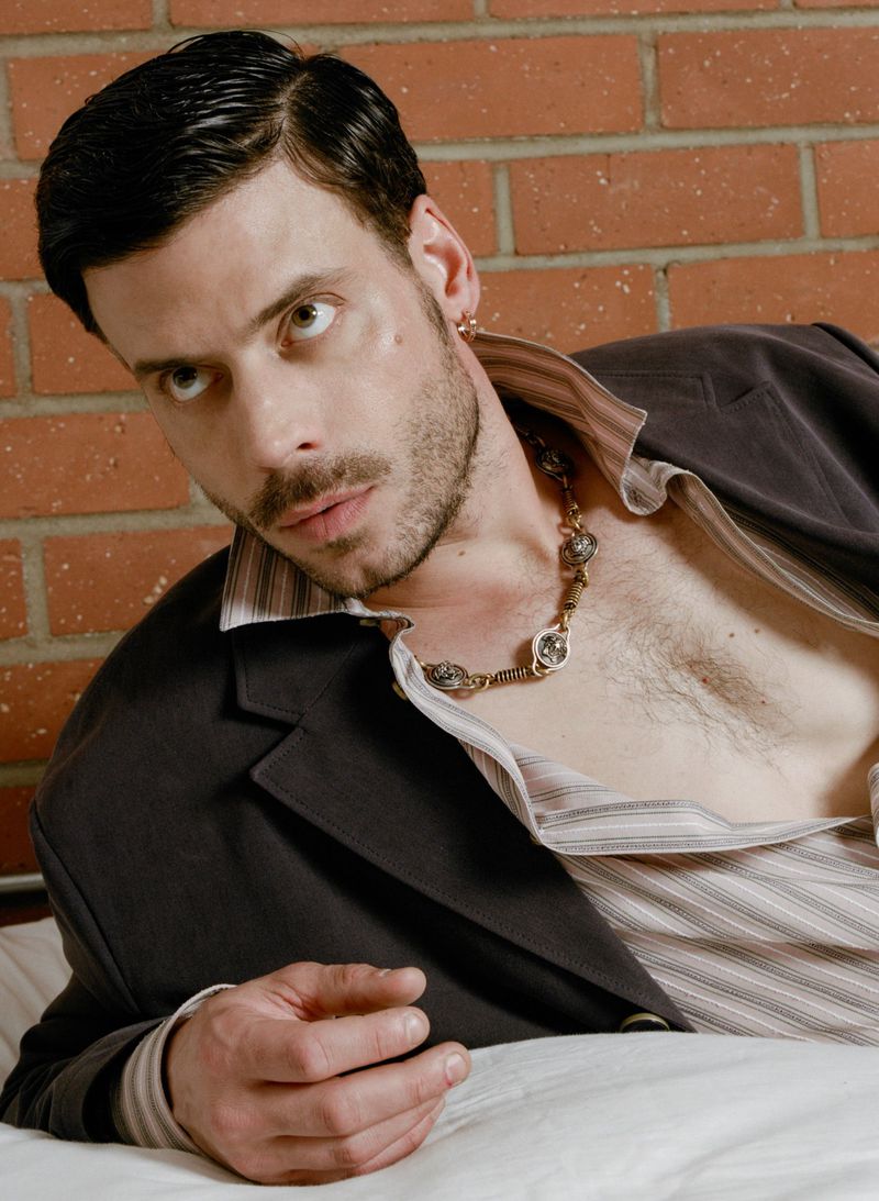 Man with dark hair and stubble in a suit jacket poses against a brick wall while laying on a white bed, gazing at the camera.