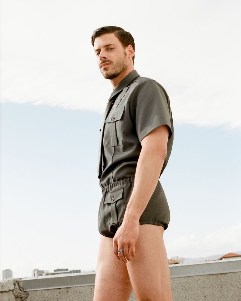 Portrait of a man in a gray playsuit on a rooftop against a bright sky. He gazes directly at the camera with a neutral facial expression.