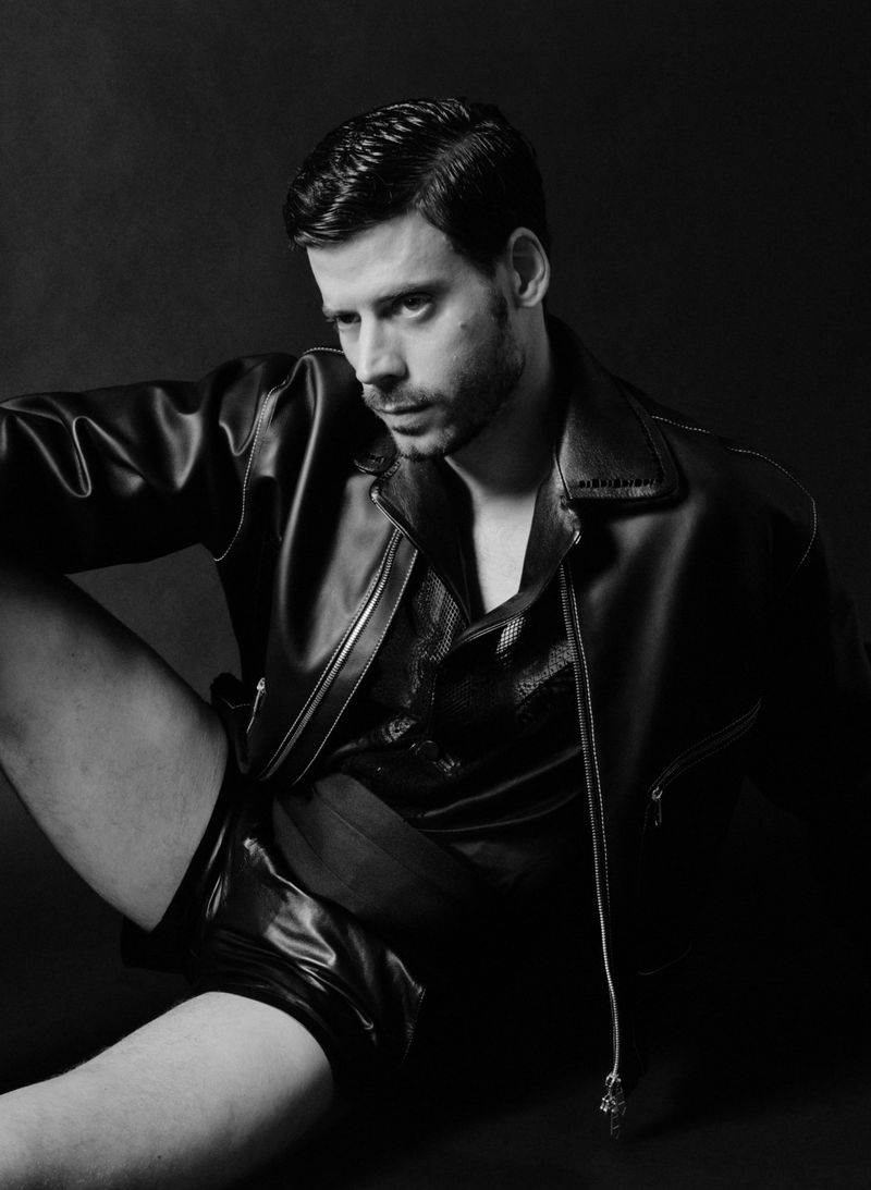Portrait of a man in a leather jacket against a dark backdrop. He is sitting and looking to the side with a neutral expression.