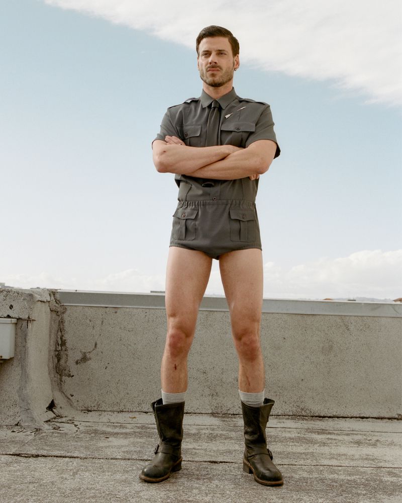 Man in playful fashion pose wearing a gray short-sleeved top, briefs, boots, and socks, standing on a rooftop against a blue sky.