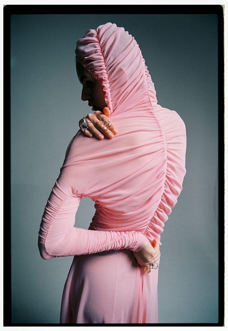 Woman in a pink hooded dress with gathers at the back and wrists, posed against a soft gray background in a studio environment.