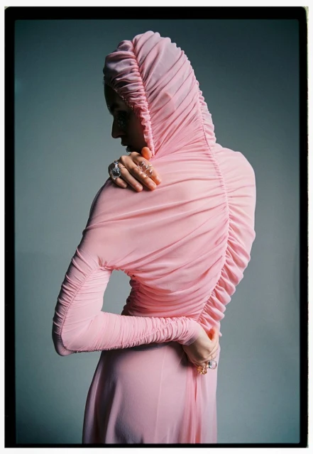 Woman in a pink hooded dress with gathers at the back and wrists, posed against a soft gray background in a studio environment.