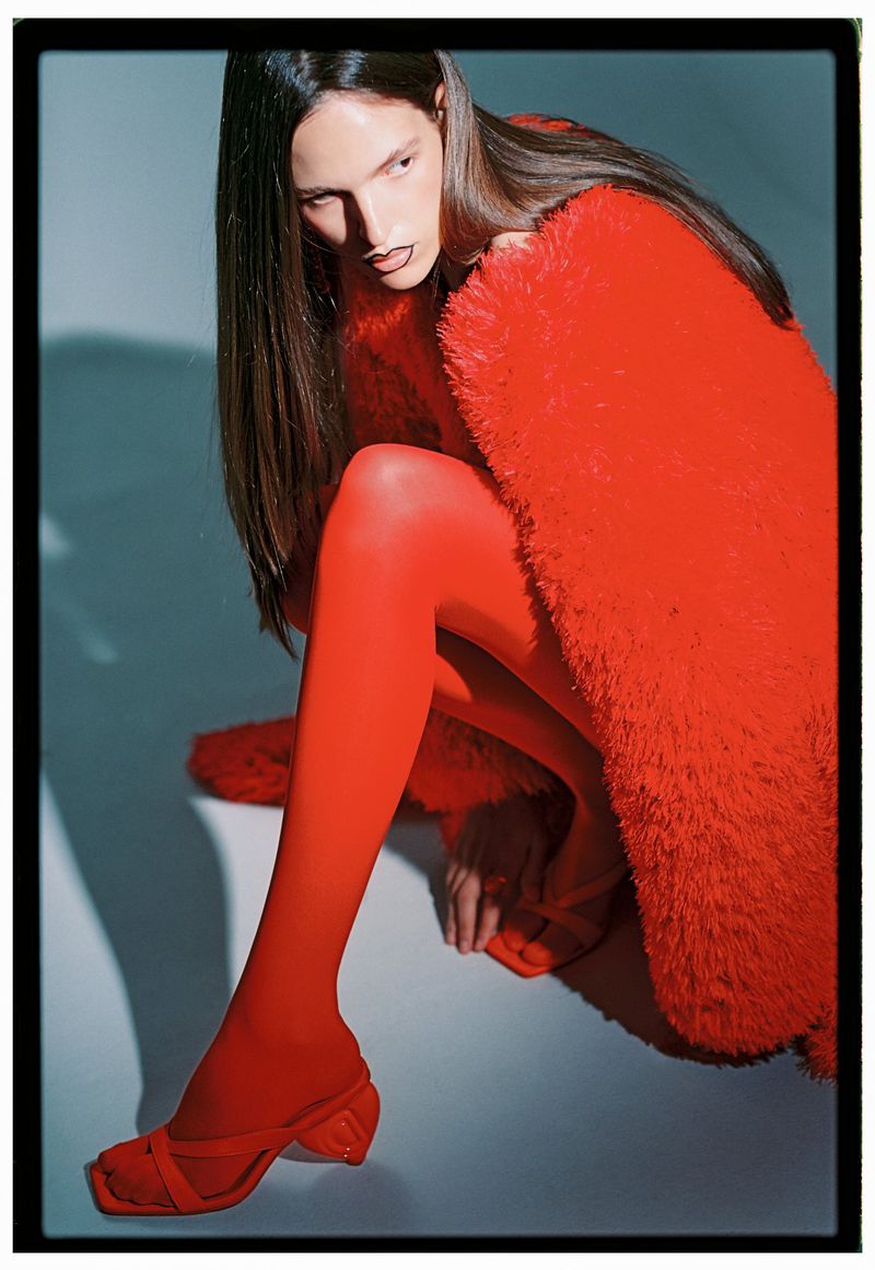 A fashionable woman in red tights, heels, and a fur coat poses in a studio, showcasing a bold and striking monochromatic fashion statement.