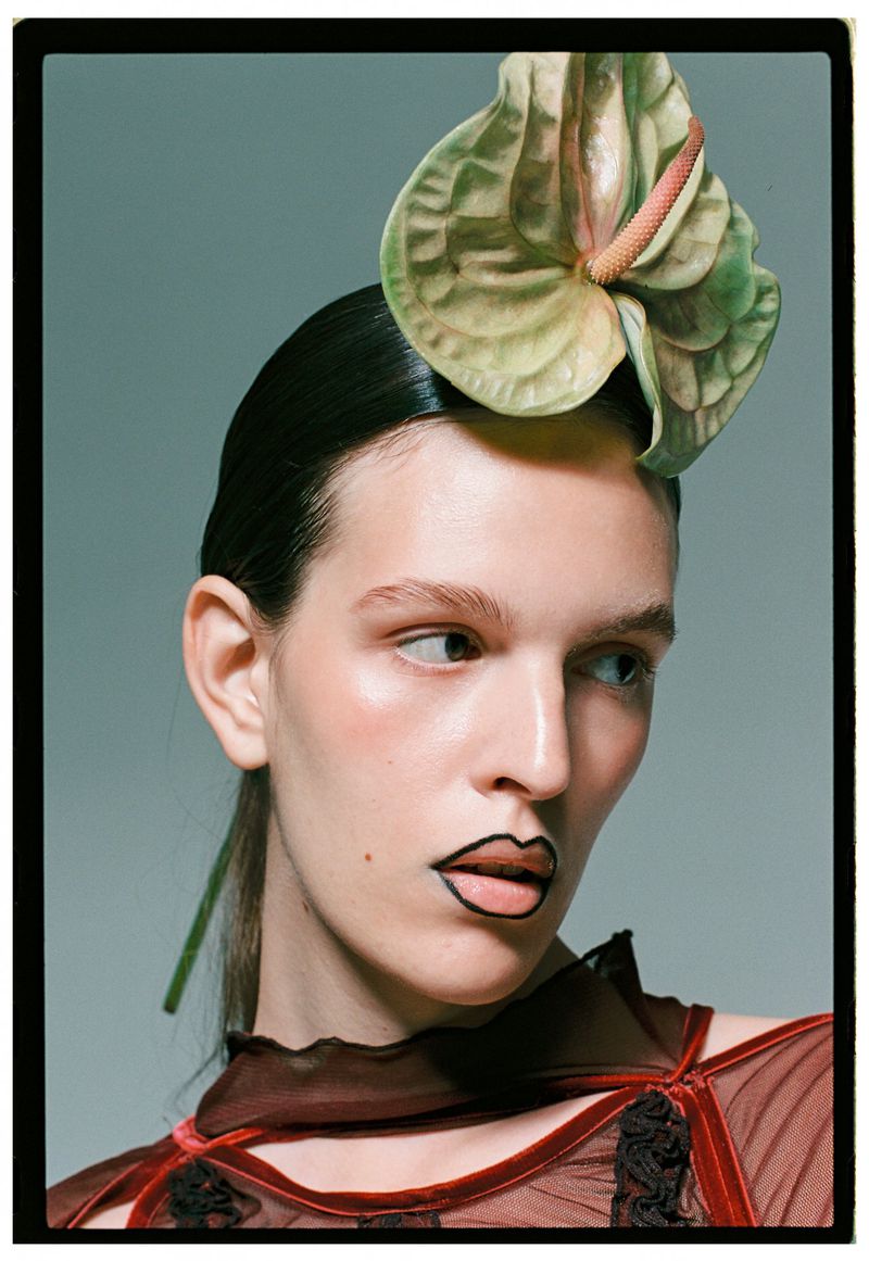 Close-up portrait of a woman with slicked-back dark hair, wearing a sheer top, and a large anthurium flower as a headpiece.