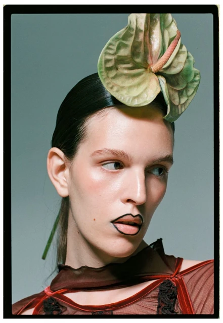 Close-up portrait of a woman with slicked-back dark hair, wearing a sheer top, and a large anthurium flower as a headpiece.