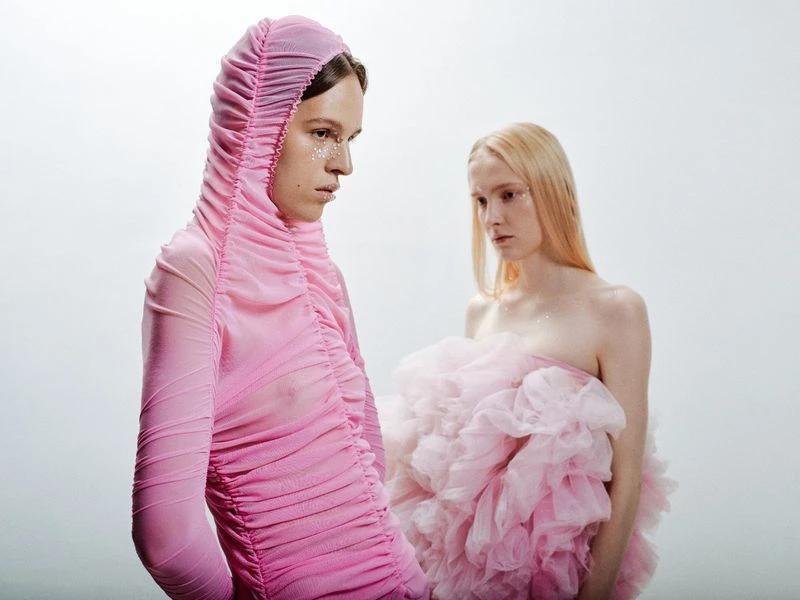 Two fair-skinned adults in a studio setting with pink tulle garments and light makeup, set against a pale backdrop, and lit with soft light.