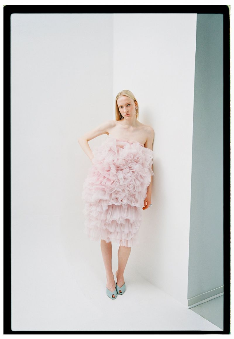 A blonde woman poses in a pink, layered ruffle dress against a white corner, complemented by light blue heels in a fashion editorial.