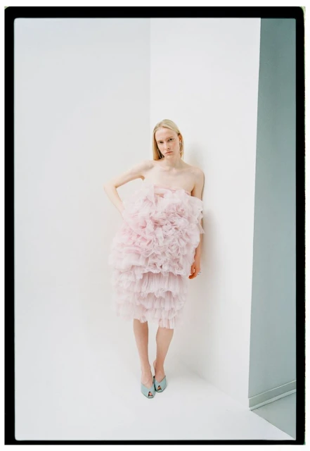 A blonde woman poses in a pink, layered ruffle dress against a white corner, complemented by light blue heels in a fashion editorial.