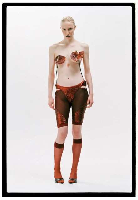 A pale-skinned woman with dark lipstick stands in a studio wearing a fashion outfit of red anthirium flowers and red sparkling bottoms.