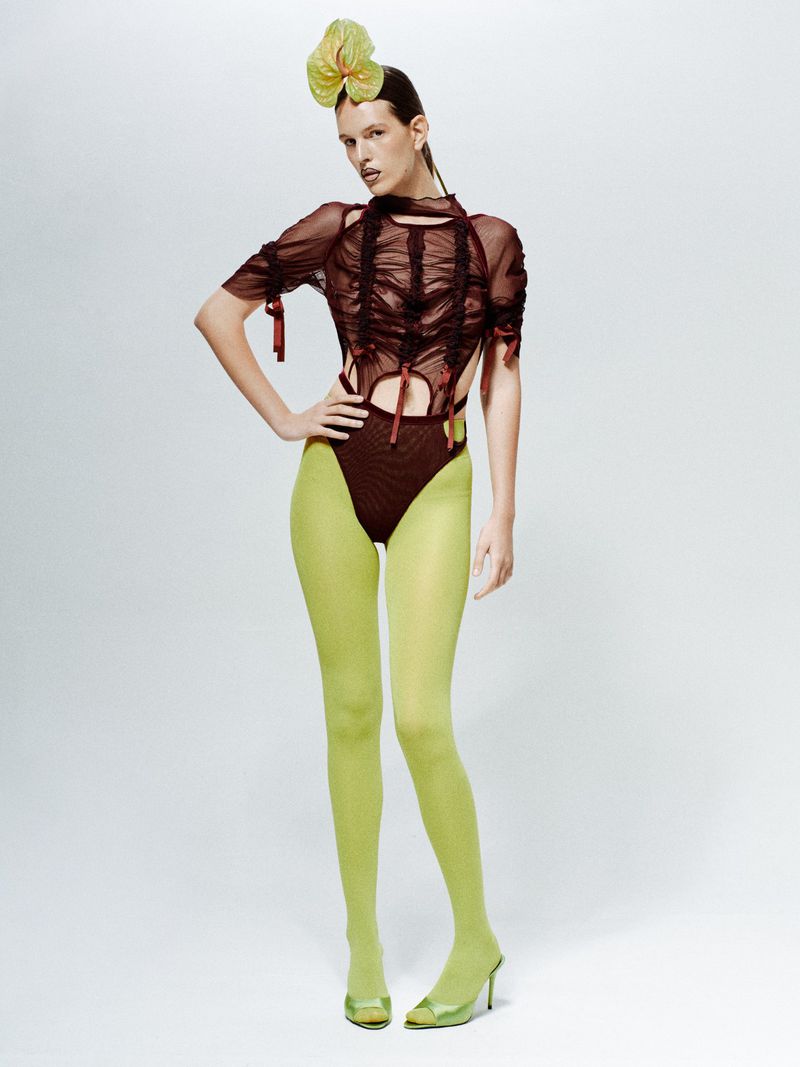 Woman poses in sheer top and green tights in a studio. Her hair is styled back, and she is wearing a green flower hairpiece.