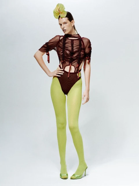 Woman poses in sheer top and green tights in a studio. Her hair is styled back, and she is wearing a green flower hairpiece.