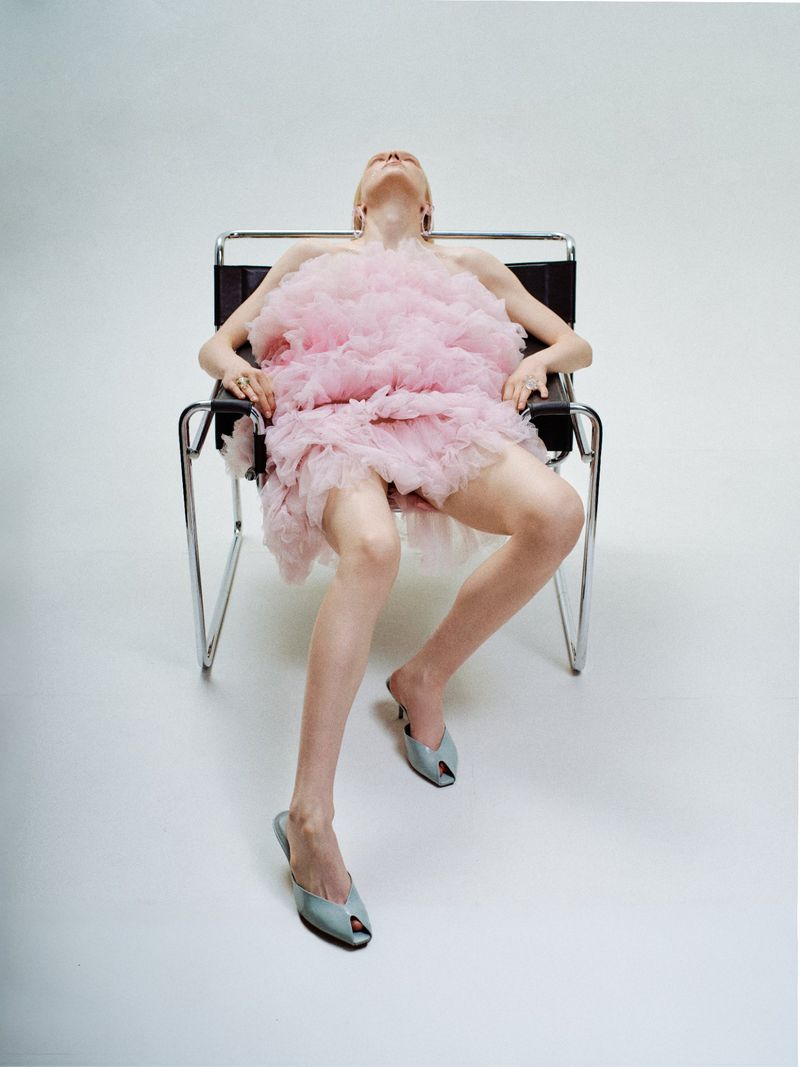 A young adult female is seated in a modern chair in a photo studio, wearing a pink dress and stylish shoes. Full body shot.